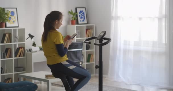 Tonårstjej tränar med exercycle i hemmet och använder sociala nät i smartphone, internetberoende av moderna ungdomar — Stockvideo