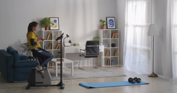 Mujer está haciendo deporte en la sala de estar de su apartamento moderno, sentado en bicicleta estática y el uso de teléfonos inteligentes, estilo de vida saludable — Vídeos de Stock