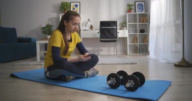 Genç kadın dambıllarıyla spor minderinde dinleniyor, internet sörfü için akıllı telefon kullanıyor, evde egzersiz yapıyor.