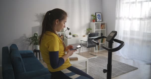 Дівчина-підліток читає повідомлення та новини в соціальних мережах за допомогою клітини, сидячи на стаціонарному велосипеді вдома, спортивне тренування для шолома — стокове відео