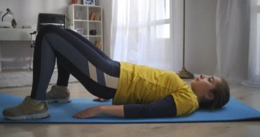 Evde sabah antrenmanı, genç kız tek başına oturma odasında antrenman yapıyor, yerde yatıyor ve kalçalarını kaldırıyor, sağlıklı bir yaşam tarzı.