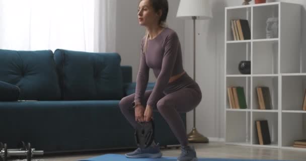 Ćwiczenia fitness w domu dla dobrej kondycji ciała, młoda wysportowana kobieta trenuje w salonie, robi przysiady z ciężarem w rękach — Wideo stockowe
