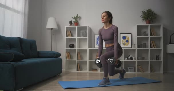 Atletik kadın hafta sonları oturma odasında antreman yapıyor, ellerinde dambıllarla, bacaklarında ve kalçalarında çalışan kaslarıyla çömeliyor. — Stok video