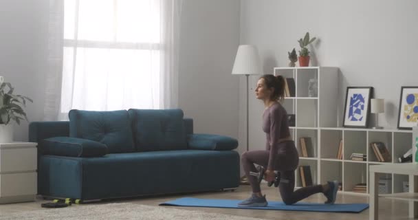 Домогосподарка тренується вдома, робить вправи для сідниць і ніг, присідаючи гантелями в руках, зберігаючи пристосованість і втрачаючи вагу — стокове відео