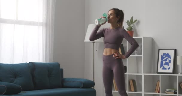 Mujer atlética cansada joven está bebiendo agua después del entrenamiento de fitness en casa, retrato medio en la sala de estar, relajarse y descansar — Vídeo de stock