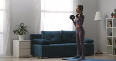 Günlük ev antrenmanı, genç atletik kadın gündüz vakti oturma odasında dambıllarla antrenman yapıyor, modern iç mekanlarda sporcu portresi.