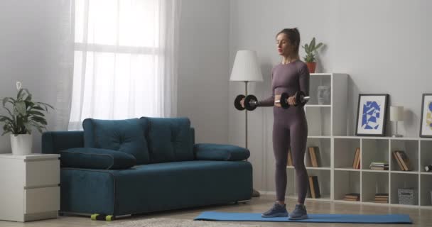 Joven mujer deportiva está entrenando con pesas, de pie en la sala de estar, retrato de la deportista en el interior moderno — Vídeos de Stock