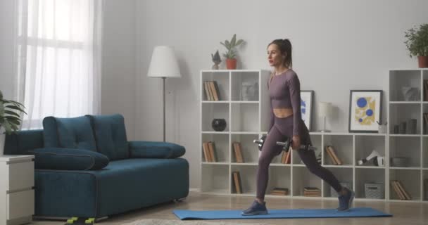 Довольно стройная женщина делает упражнения для ног дома, приседает с гантелями в руках, заботясь о хорошей форме тела — стоковое видео
