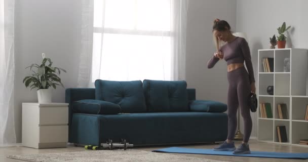 Sportieve vrouw is kijken op fitness tracker en doen hurken met gewicht in appartement, home fitness activiteit — Stockvideo