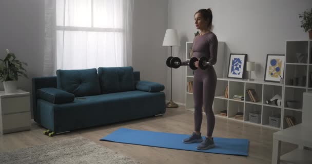 Mujer delgada está levantando pesas en la sala de estar, el entrenamiento de bíceps en casa para una buena forma del cuerpo, el entrenamiento diario en casa en el apartamento — Vídeos de Stock