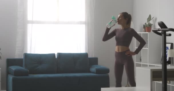 Sportieve vrouw is het drinken van water tijdens de pauze thuis workout, ontspannen in de woonkamer op zonnige dag, sport activiteit — Stockvideo
