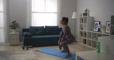 Evde ağırlıklarla antrenman yapan genç sporcu kadın, ellerinin ağırlığıyla, kalçalarının ve bacaklarının kaslarıyla çömelme hareketleri yapıyor.