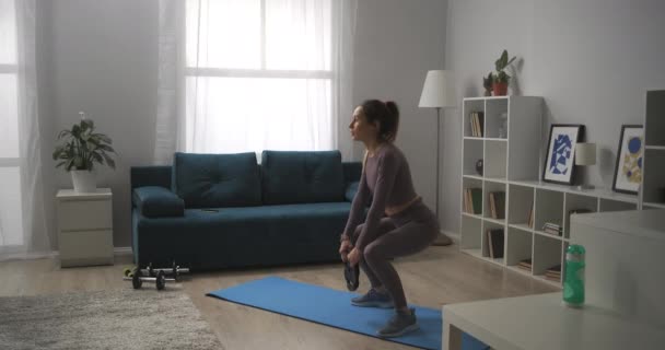Тренування з вагами вдома, молода спортсменка робить присідання з вагою в руках, працюючи м'язи сідниць і ніг — стокове відео