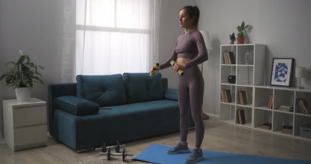 Sportos nő egyedül edz a nappaliban, otthoni fitnesz gyakorlatok súlyzókkal, edzés a lakásban, fittség fenntartása — Stock videók