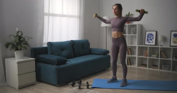 Mujer sonriente vestida ropa deportiva está entrenando con pequeñas mancuernas en la sala de estar, entrenamiento en casa para una buena forma del cuerpo — Vídeos de Stock