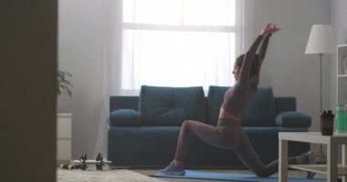 Evde yoga antrenmanı, genç sporcu kadın yalnız başına oturma odasında antrenman yapıyor, hafta sonları antrenman yapıyor, savaşçı pozu veriyor.