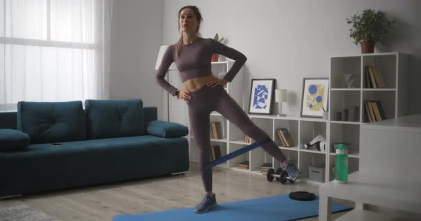 Спортивная женщина тренируется с фитнес упругой лентой, напрягая мышцы ног для потери веса и хорошей формы, домашняя тренировка — стоковое видео
