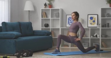 Evde egzersiz yaparken, genç bir kadın tek başına oturma odasında antrenman yapıyor, modern bir dairede paspasın üzerinde oturuyor.