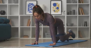Bacak kasları için elastik bantla spor yapıyor, kadın oturma odasında el ve dizlerinin üzerinde duruyor ve bacaklarını yukarı hareket ettiriyor.