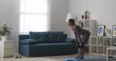 İnce fitness modeli, elinde dambıllar, modern dairenin oturma odasında duruyor, genç bir kadının güç eğitimini alıyor.