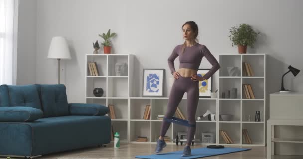 Entrenamiento aeróbico y gimnasia en el hogar, la mujer está moviendo las piernas de pie en la sala de estar, cuerpo femenino deportivo vestida ropa deportiva de moda — Vídeo de stock