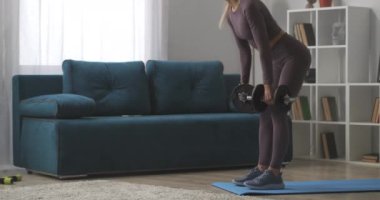 Genç sarışın, dambıllarla antrenman yapıyor, oturma odasında eğiliyor, evde spor yapıyor, sağlıklı bir yaşam sürüyor.