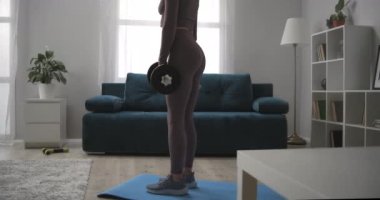 Güzel bir kadın, elinde dambıllarla antrenman yapıyor, ağırlıkla öne eğiliyor, oturma odasında egzersiz yapıyor, sağlıklı bir yaşam sürüyor.