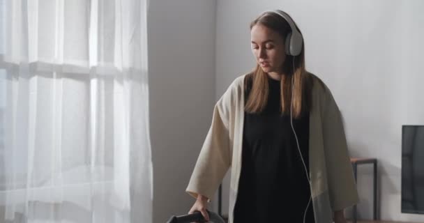 Молода жінка використовує пуф для прибирання вдома, слухаючи пісню з навушниками та весело танцюючи у вітальні — стокове відео