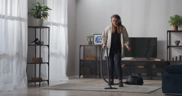 Wanita muda ceria membersihkan karpet dengan vacuum cleaner di kamar dan menari, mendengarkan musik dengan headphone — Stok Video