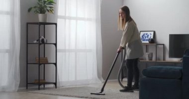 Genç bayan modern dairenin oturma odasında halı süpürüyor, elektrikli süpürgeyle odada duruyor.