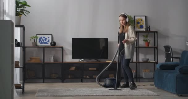 Jonge vrouw met hoofdtelefoon luistert naar muziek tijdens huishoudelijk werk, stofzuigen tapijt en dansen, luisteren naar zang en zingen — Stockvideo