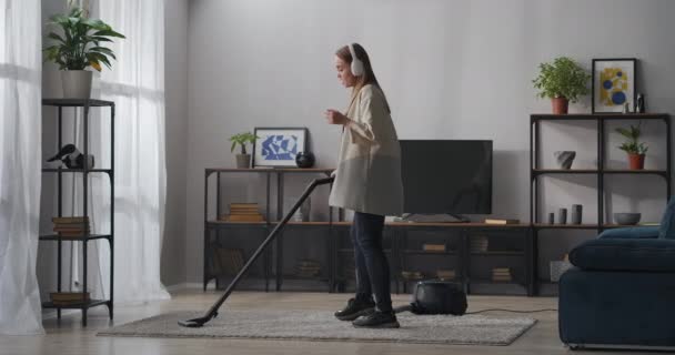 Alegre joven mujer está escuchando música por los auriculares y alfombra de limpieza por aspiradora en la habitación — Vídeo de stock