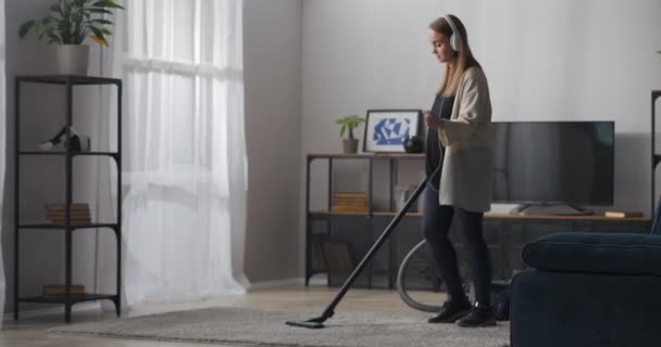 Jonge mooie vrouw is stofzuigen in appartement op zonnige dag, het luisteren naar muziek door een koptelefoon en dansen — Stockvideo