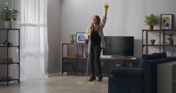 Ama de casa joven está bailando con fregona en el apartamento, cantando canción y escuchando música, lavando piso en la sala de estar — Vídeos de Stock