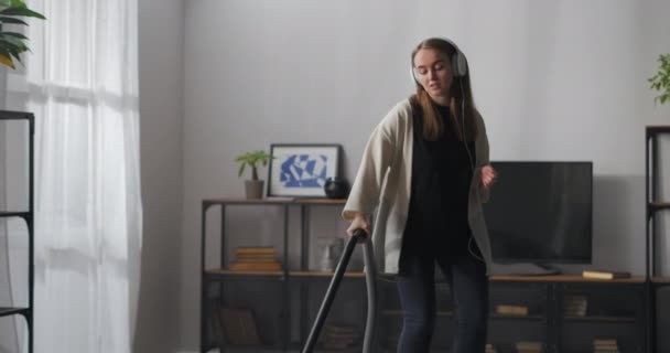 Vrolijke vrouw doet schoonmaken in de kamer, stofzuigen vloer door moderne stofzuiger, luisteren naar muziek en dansen — Stockvideo