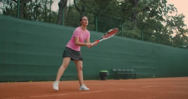 Μια νεαρή γυναίκα προπονείται σε μια σκληρή επιφάνεια γήπεδο τένις το ηλιοβασίλεμα, περιμένοντας να εξυπηρετήσει, Forehand, πλήρη βολή — Αρχείο Βίντεο