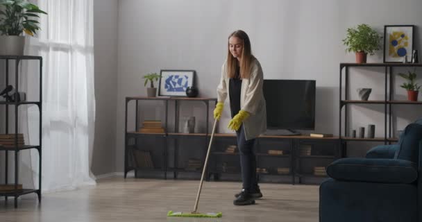 Jonge blonde vrouw is wassen vloer in appartement op zonnige dag, met behulp van dweil en rubberen handschoenen, huishouding — Stockvideo