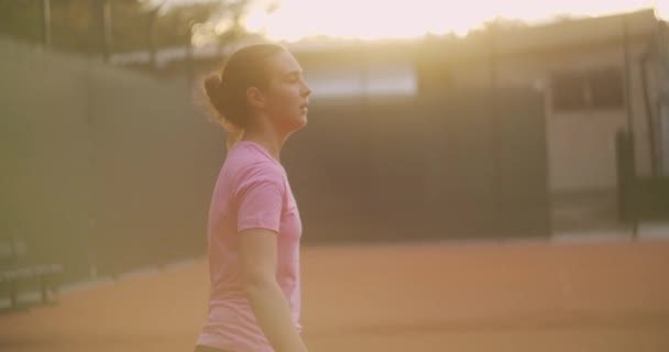 A menina está cansada durante o treinamento de tênis em um campo aberto com uma superfície de sujeira. lentamente se aproxima da bola e bate com um backhand — Vídeo de Stock
