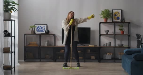 Ung kvinna sjunger i mopp som i mikrofon, tvättgolv och dansar med mopp i lägenhet, bra och glad stämning, städning och städning — Stockvideo