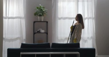 Neşeli kadın paspasla dans ediyor ve oturma odasında şarkı söylüyor, evi temizliyor ve keyfi yerinde, ev işi ve temizlik yapıyor.