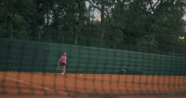 Tenis antrenmanındaki bir kadın önce ezme topu, sonra da ters vuruş yapar. Gün batımında dışarıda antrenman. Yeşil çitli bir Clay kortu. Geniş açı, ağ boyunca..