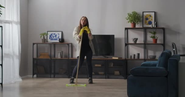 Limpieza en la sala de estar, la mujer está cantando en fregona como en el micrófono, lavando piso y bailando en apartamento — Vídeo de stock