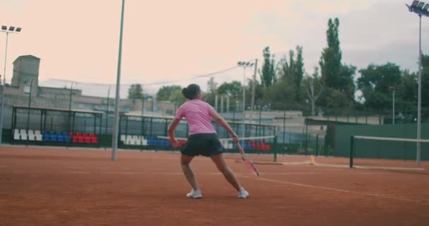 Ung kvinna går genom tennisbanan med oväsen. Baksidan av attraktiv brunett kvinna i rosa skjorta och svarta shorts in tennis hardcourt. Full längd följ skott med kopieringsutrymme — Stockvideo