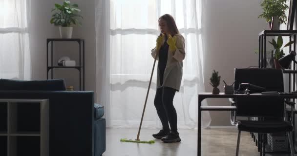 Femme de ménage lave le sol dans le salon de l'appartement, danse avec la serpillière et le chant, écoute de la musique — Video