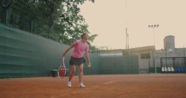 Kız servis yapmadan önce duruşunu aldı. Smash, zıpladıktan sonra, sağ eliyle tenis topuna vuruyor, yeşil çitli bir Clay kortu. Geniş açı, ağ boyunca..