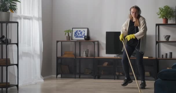 Mulher engraçada com fones de ouvido é lavar o chão e ouvir música, limpeza da sala de estar, tarefas domésticas — Vídeo de Stock