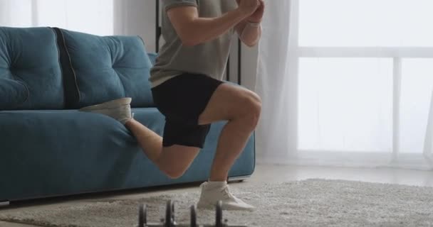 Pelatihan rumah dari olahragawan muda, latihan di ruang tamu, melakukan jongkok dengan satu kaki, menggunakan sofa seperti peralatan olahraga — Stok Video