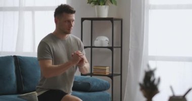 Sporcu formda, oturma odasında antrenman yapıyor, tek bacakla çömeliyor, kaslarını geriyor, spor yapıyor.