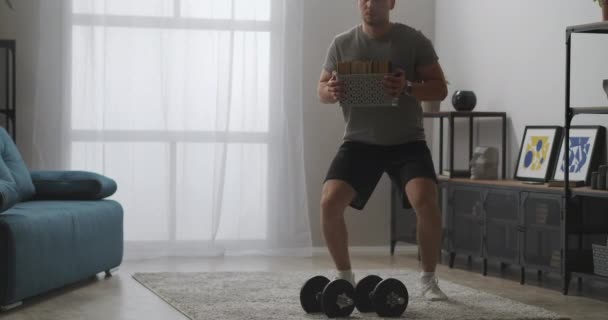 Home gym at self-isolation, handsome man is squatting with books like weight in living room, home gym and fitness — Stock Video