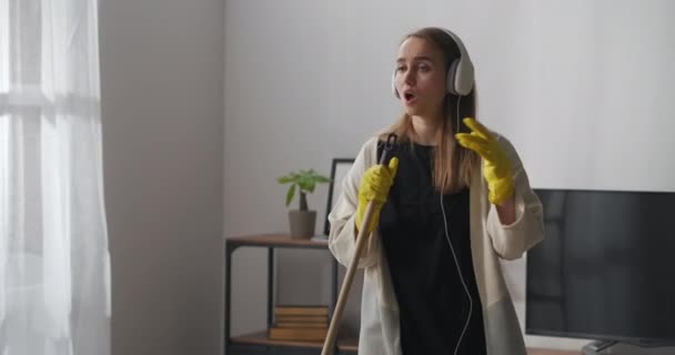 Positieve vrouw zingt in dweilen tijdens het schoonmaken in de flat, luisteren naar muziek door een koptelefoon, portret geschoten in huis — Stockvideo